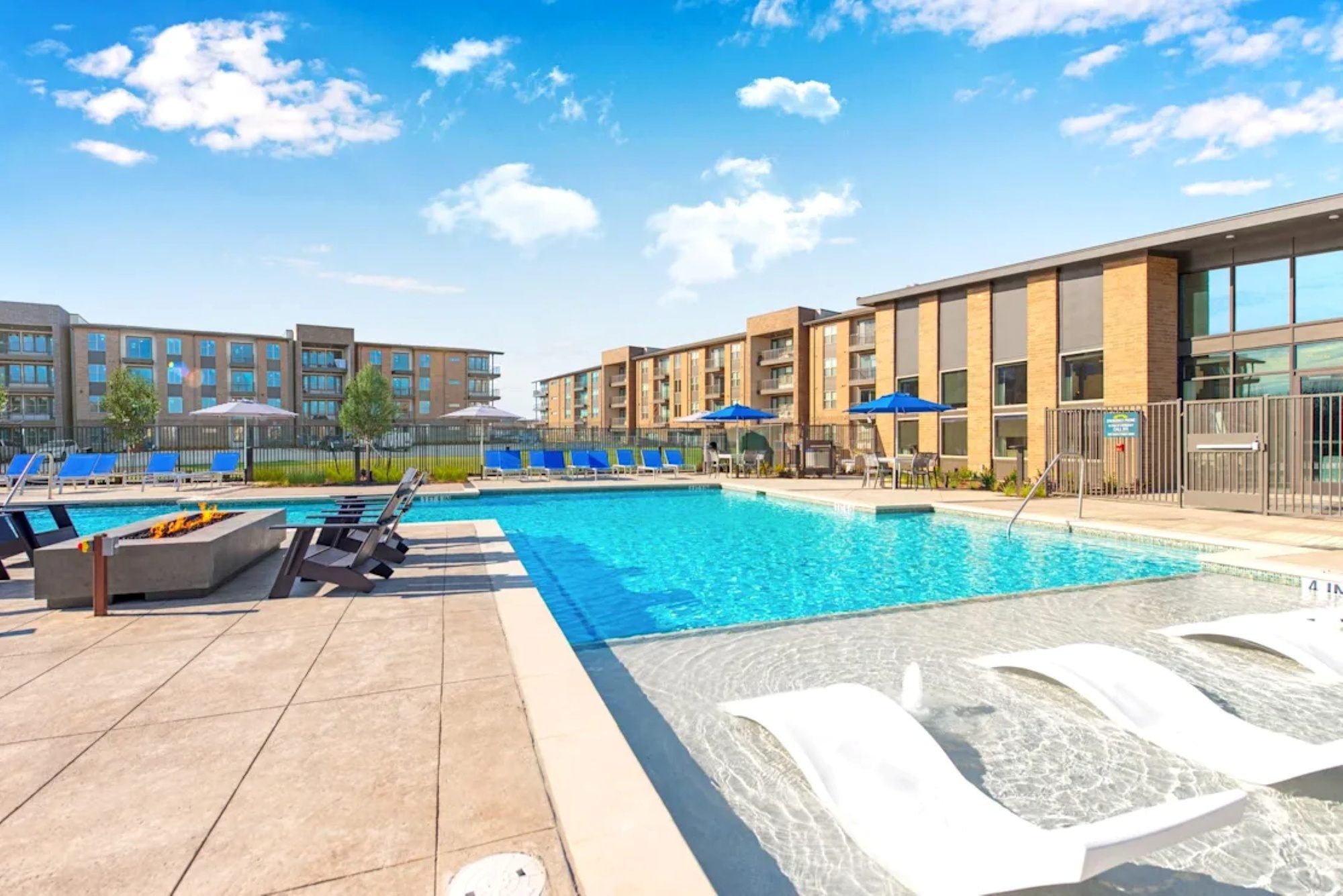 An outdoor apartment swimming pool featuring submerged white loungers, a fire pit area, and poolside seating at sunset.